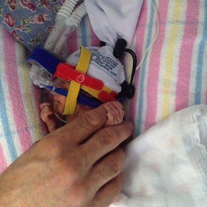 Hand holding the tiny hand of a premature baby, which is wearing a beanie and attached to tubes